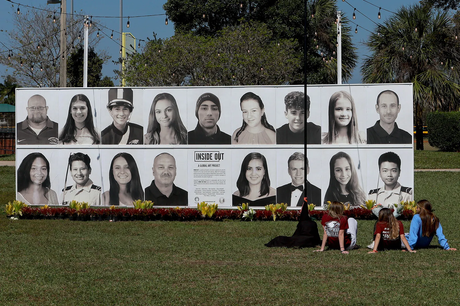 A group of people sits in front of a large poster that showcases portraits of 17 individuals, and a center graphic says "Inside Out Global Art Project".