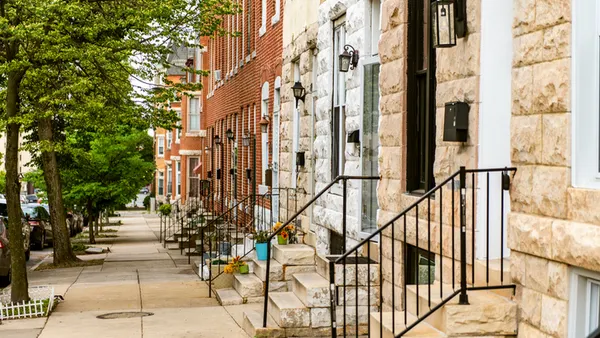 A picturesque row of urban brownstone houses with stairs and plants line a tree-shaded sidewalk.