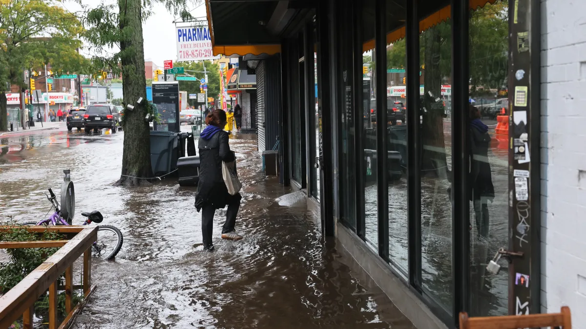 A person walks along a flooded sidewalk with a panel glass storefont to their left and a large white sign with "Pharmacy" in bold blue letters in the background and two cars driving on the far right.