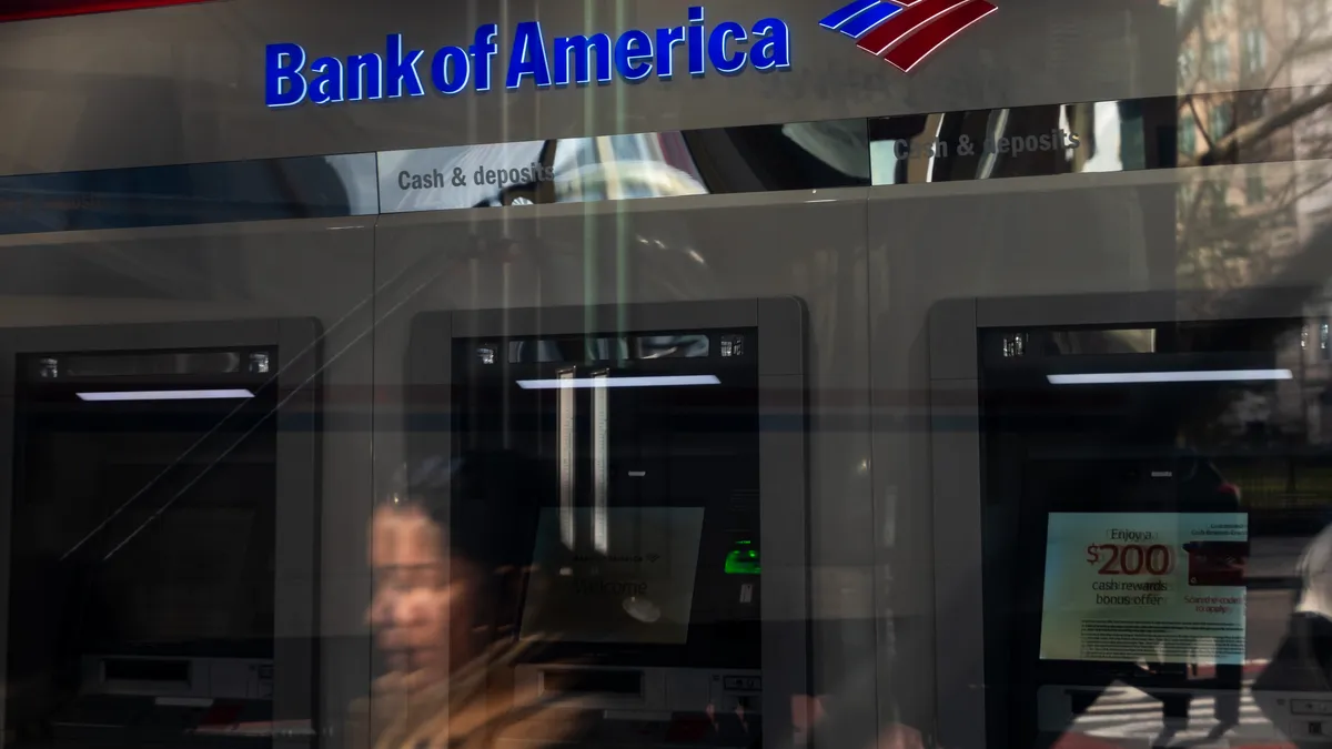 Bank of America ATMs are seen through a window. A person's reflection is seen passing by.