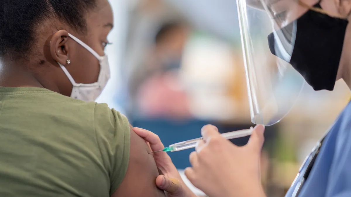 A student has their sleeve rolled up and is looking at an adult who is administering a shot into the student's arm. Both adult and student are wearing masks.