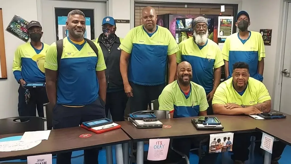 Eight people wearing high-vis shirts are standing and sitting behind a table in an office.