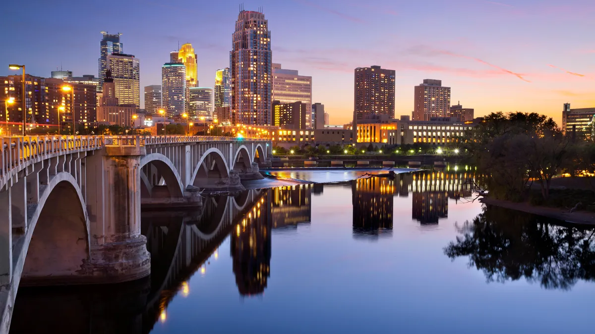 An image of the Minneapolis downtown skyline at sunset, with a bridge over a river reflecting tall buildings against a dark blue sky.