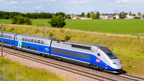 A sleek blue and silver train near a pastoral scene of greenery and small homes.