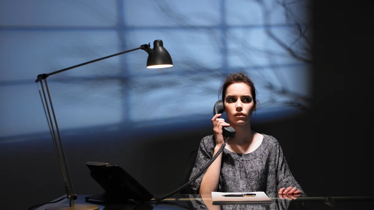 A woman sits at a desk in a dark room with a phone in her hand, looking unsettled. The shadow of a tree is projected on the wall behind her.