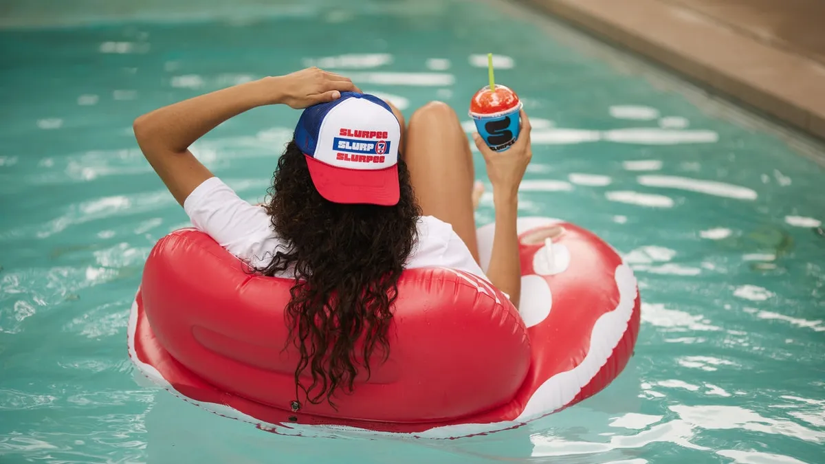 A photo of a long-haired person, whown from the back, sitting in an inflatable chair floating in a pool. They have a drink in one hand. they are wearing a red, white and blue hat that says Slurpee Slurp Slurpee.