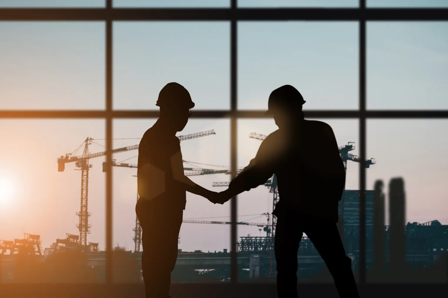 Stock image of two construction workers silhouetted against a window with construction cranes in the background.