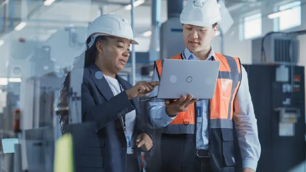 Two Professional Heavy Industry Engineers Wearing Safety Uniform and Hard Hats Discussing Factory Work on Laptop Computer