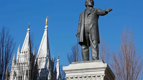 A statue of a man, Brigham Young, is seen in the foreground before the spires of a church in the background.