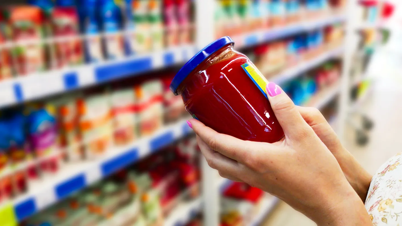 A person with pink nails holds a jar of red sauce with a blue lid in a grocery aisle. Shelves filled with colorful packaged goods are blurry in the background.