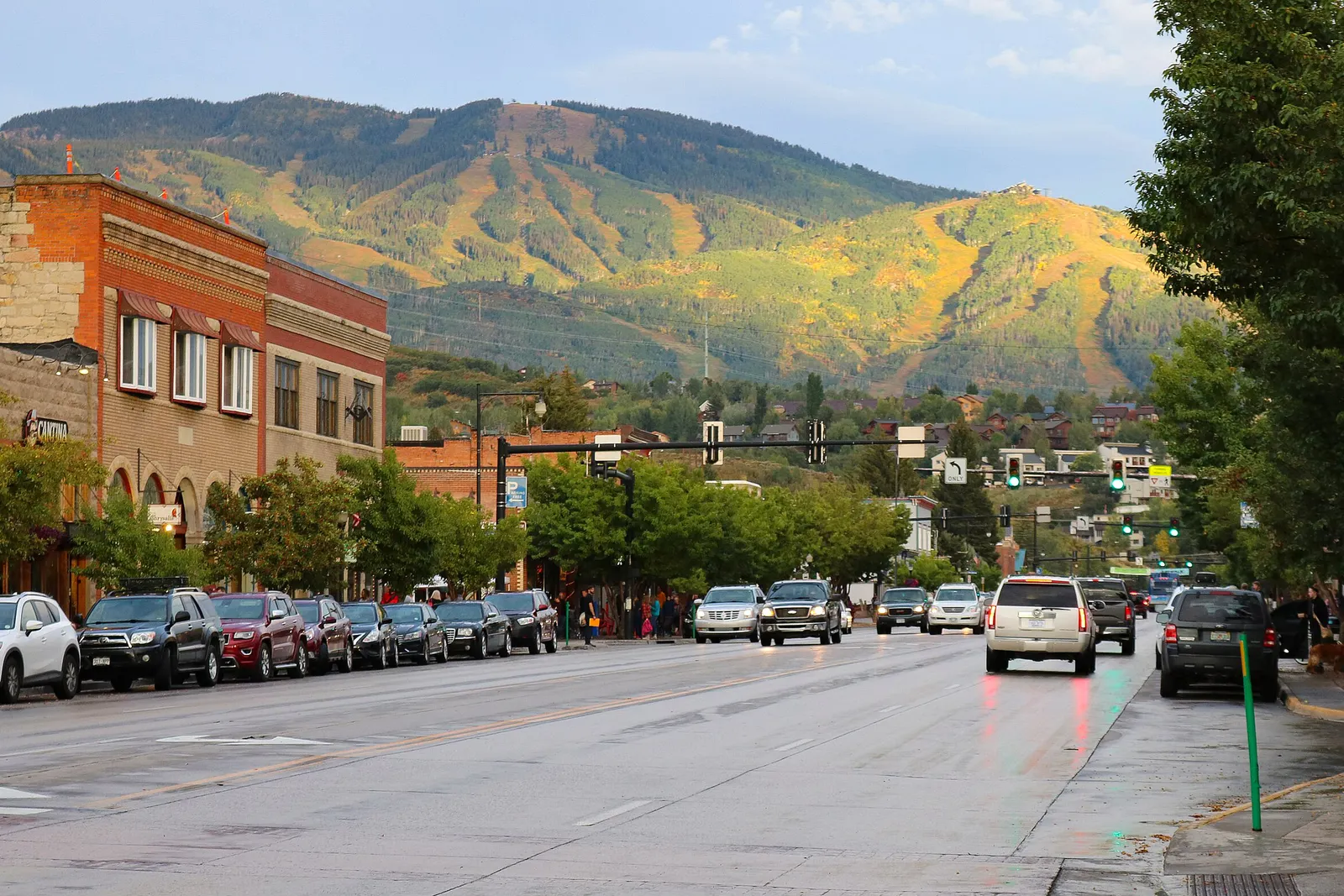 A view looking up a street in a small downtown is shown, with cars driving and parked and a mountain close in the background.