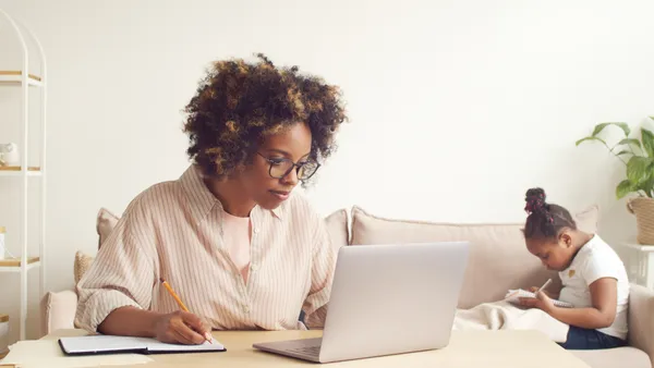 A woman works at her laptop, while a child sits on a couch in the background.