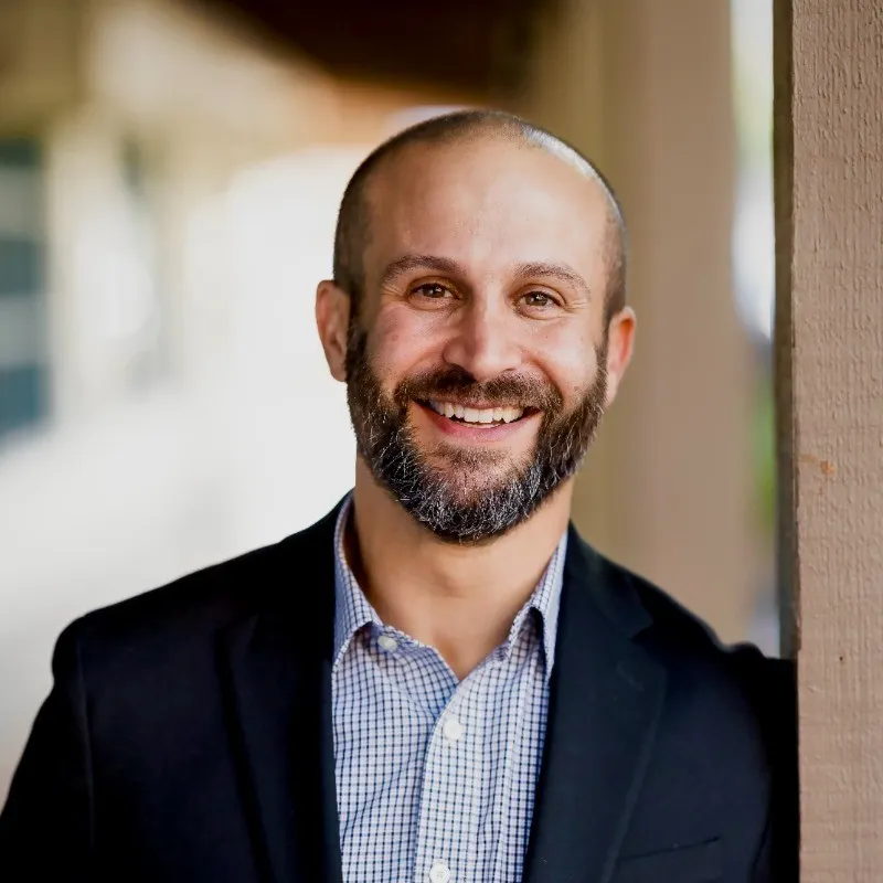 A man in a checkered button up shirt and black blazer smiling and leaning against a post.