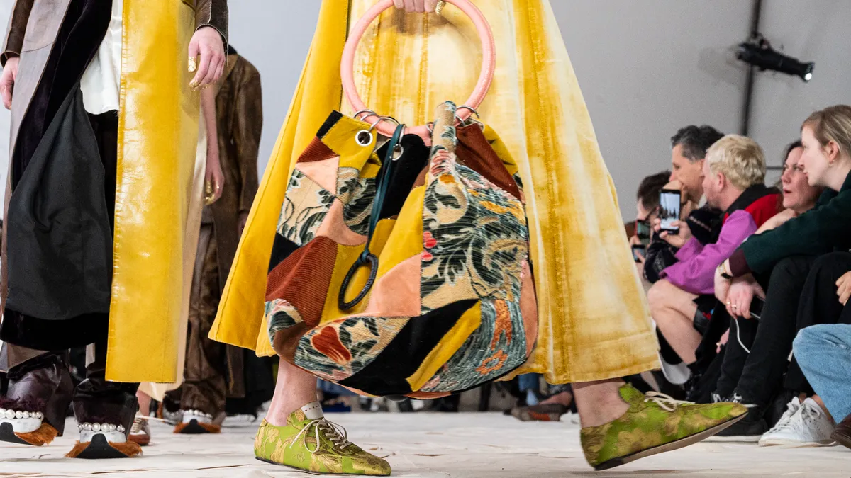 A model on a fashion runway holds a large, colorful patchwork back with a round handle.