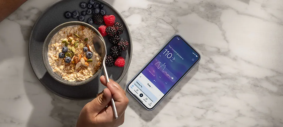 A person eats oatmeal with berries, and a phone on the counter shows their current glucose levels.