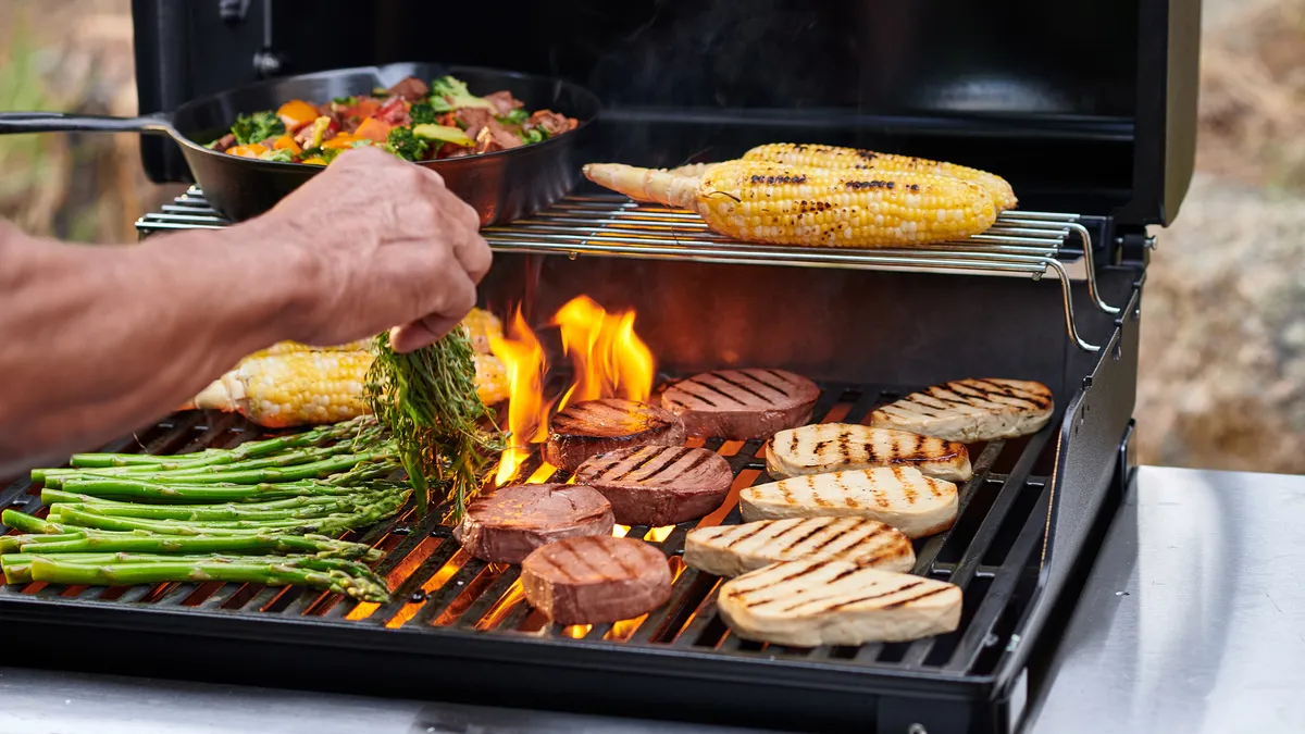 A hand holds a bundle of herbs above a fiery double-decker grill with Meati Steaks and Cutlets with grill marks on the bottom left, asparagus on the bottom right, and a frying pan of stir fry and corn