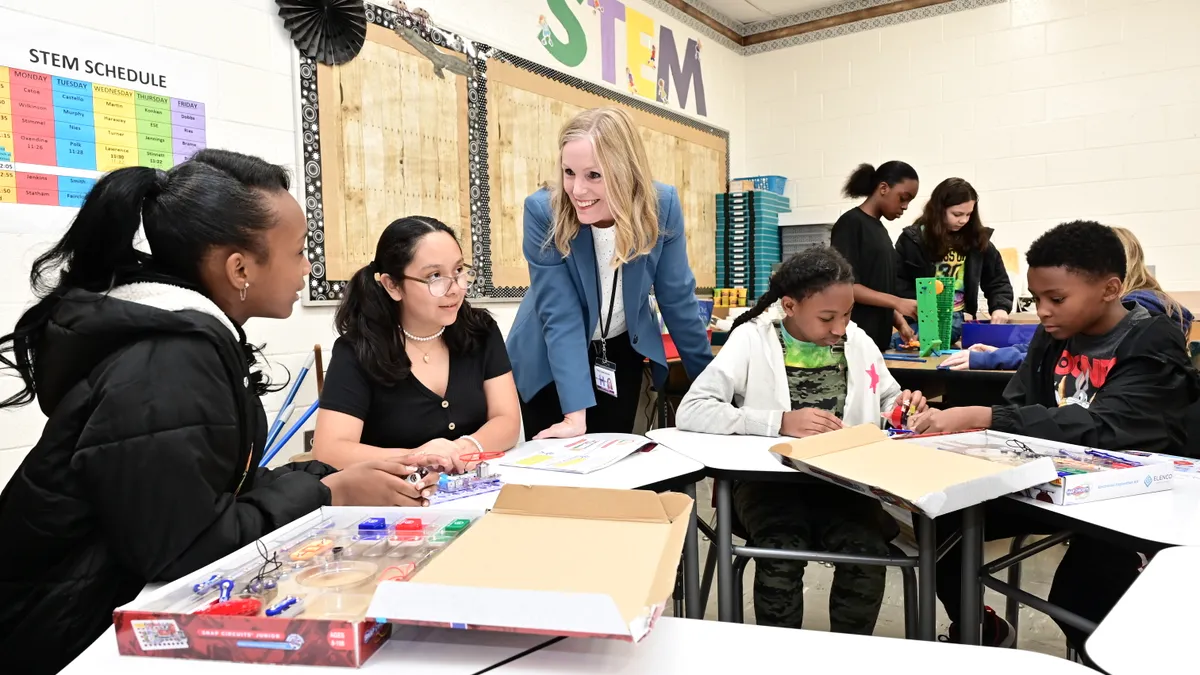 A woman superintendent interacts with a multiethnic group of students in a brightly decorated STEM classroom.