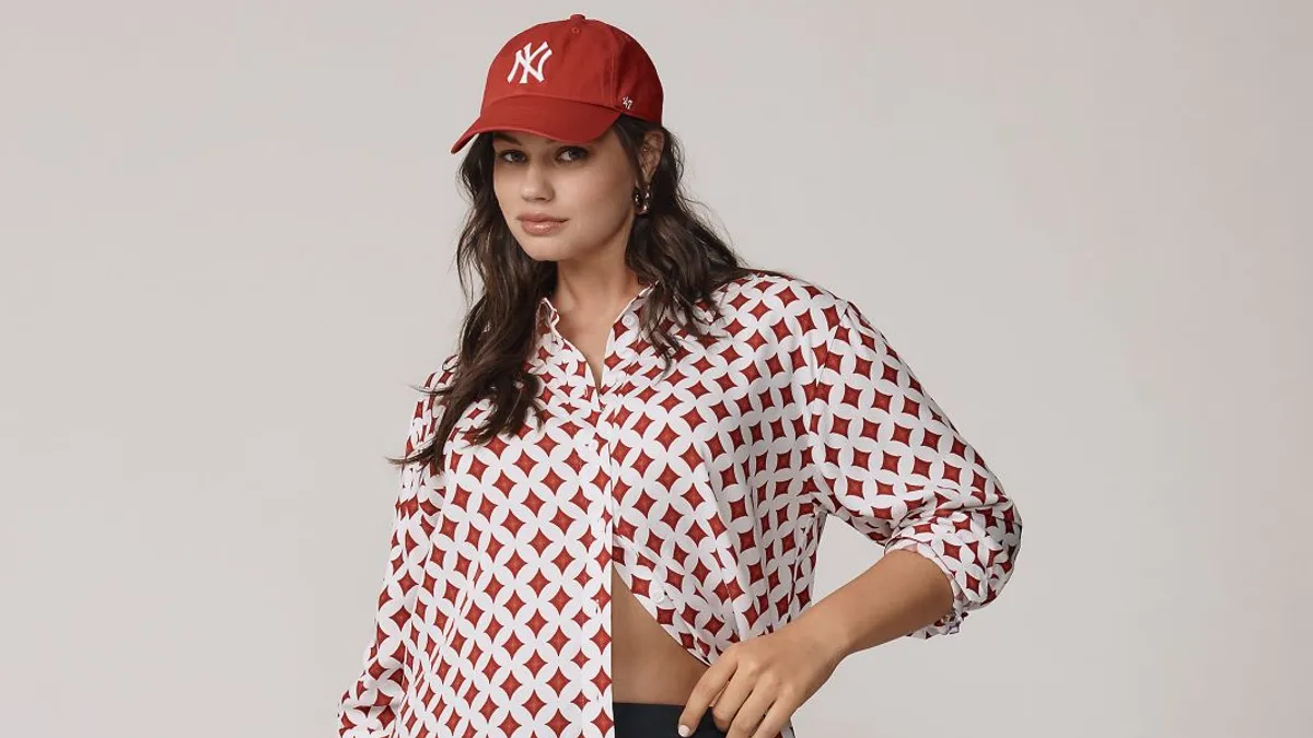 A model stands in front of a blank background wearing a skirt, button down and baseball hat.