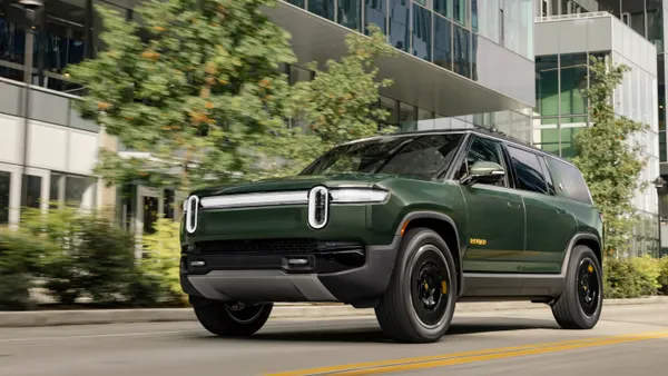A dark green Rivian R1S electric SUV driving on a city street with buildings in the background.