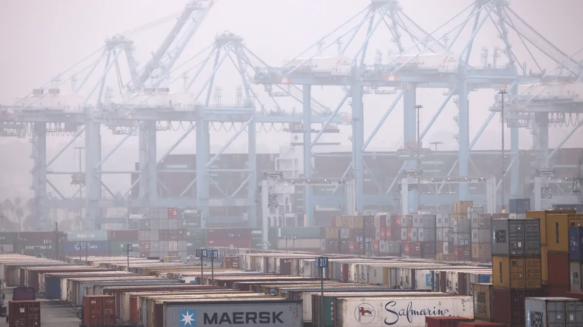 Containers are stacked at the Port of Los Angeles in front of cranes in the background.