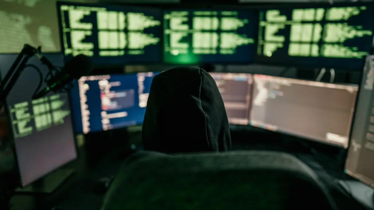 A person wearing a hoodie sits in front of multiple compluter screens with green code displayed.