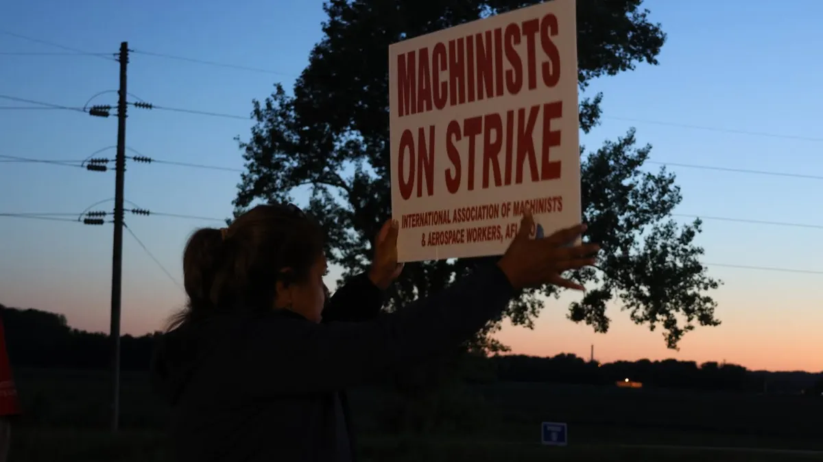 The back of a woman holding a red and white Machinists on strike sign at dusk.