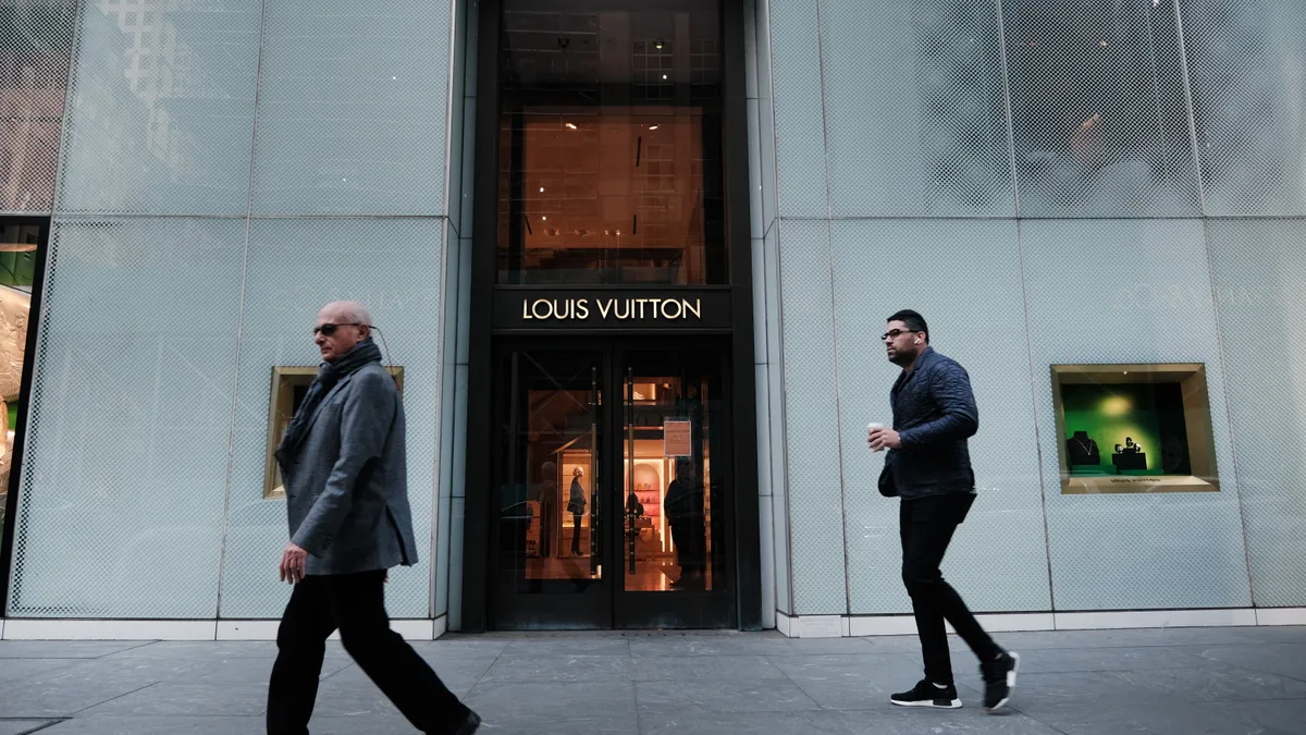 Two men in jackets and sunglasses walk past a Louis Vuitton store.