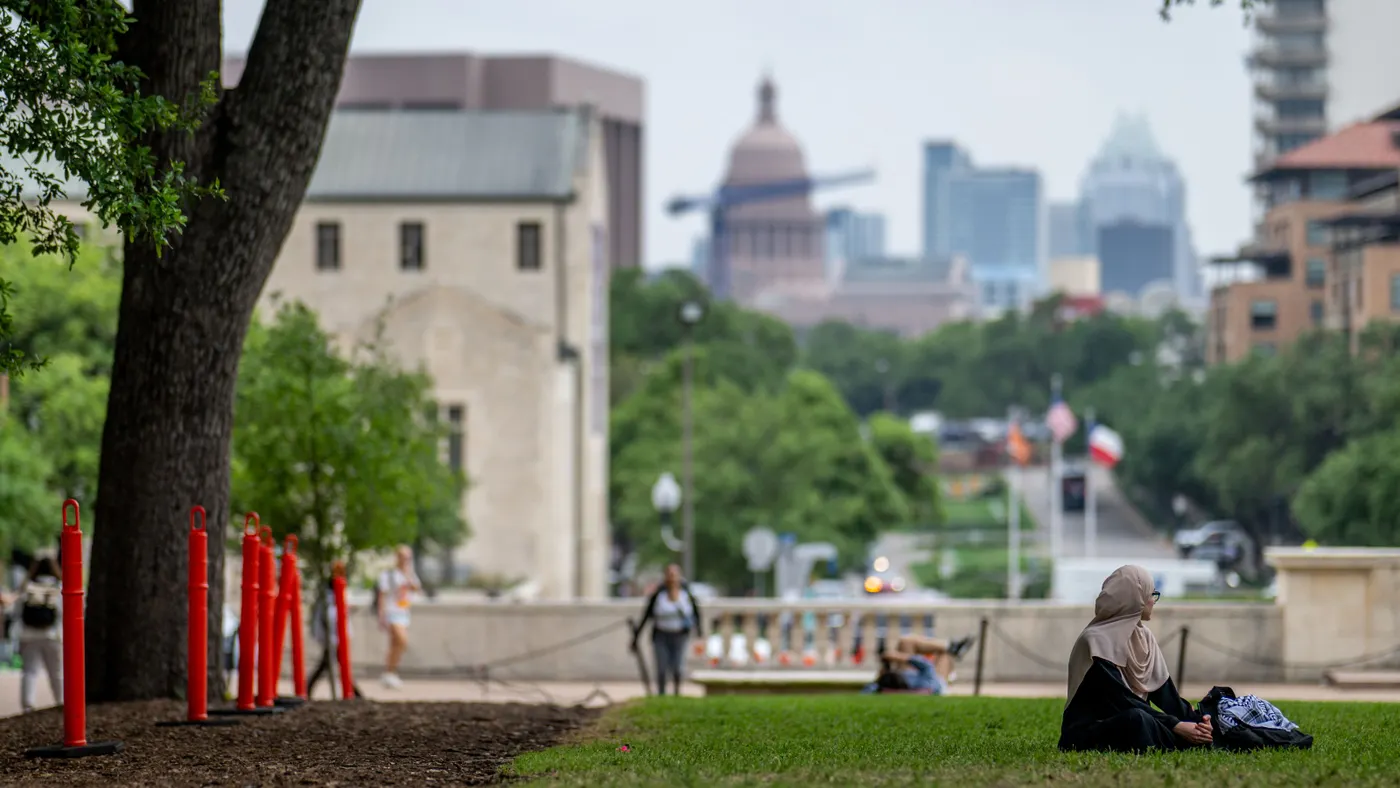 A student sits on the lawn of the University of Texas at Austin's campus with buildings in the background.
