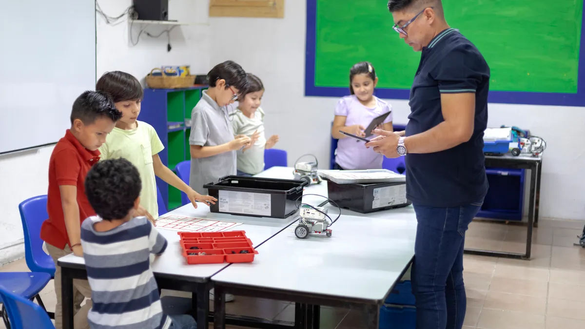 A handful of students are standing around a table in a classroom. An adult stands on the other side of the table. On the table are bins and a robot.