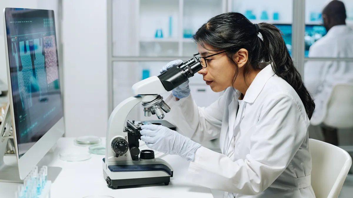 Side view of female biochemist in whitecoat, gloves and eyeglasses looking in microscope while studying virus in laboratory.