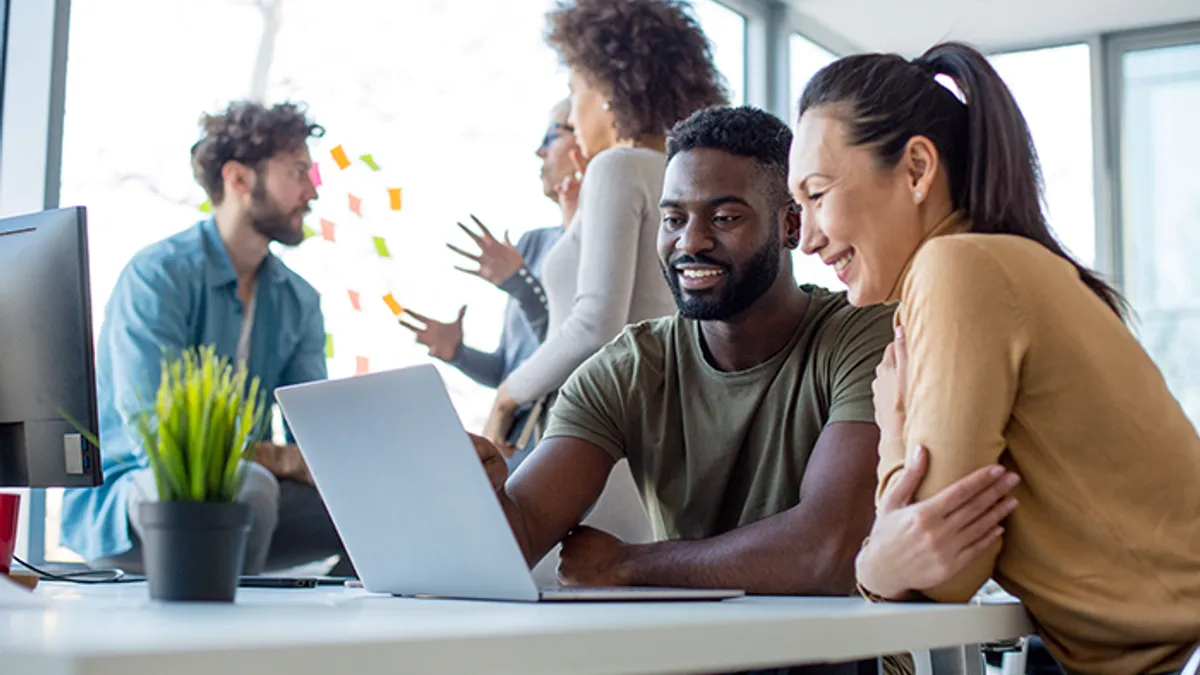 Two people smiling at laptop screen. Three people talking in background.