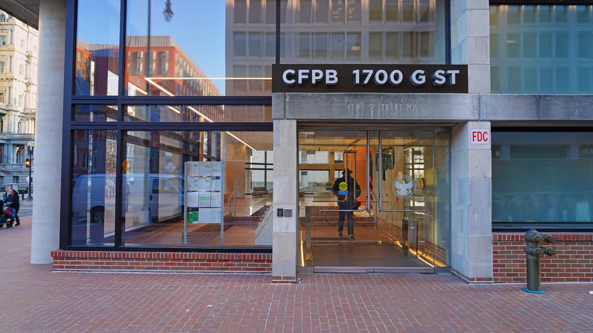 A modern cement and glass building with a sign that reads "CFPB 1700 G ST" is above a glass door entrance. 