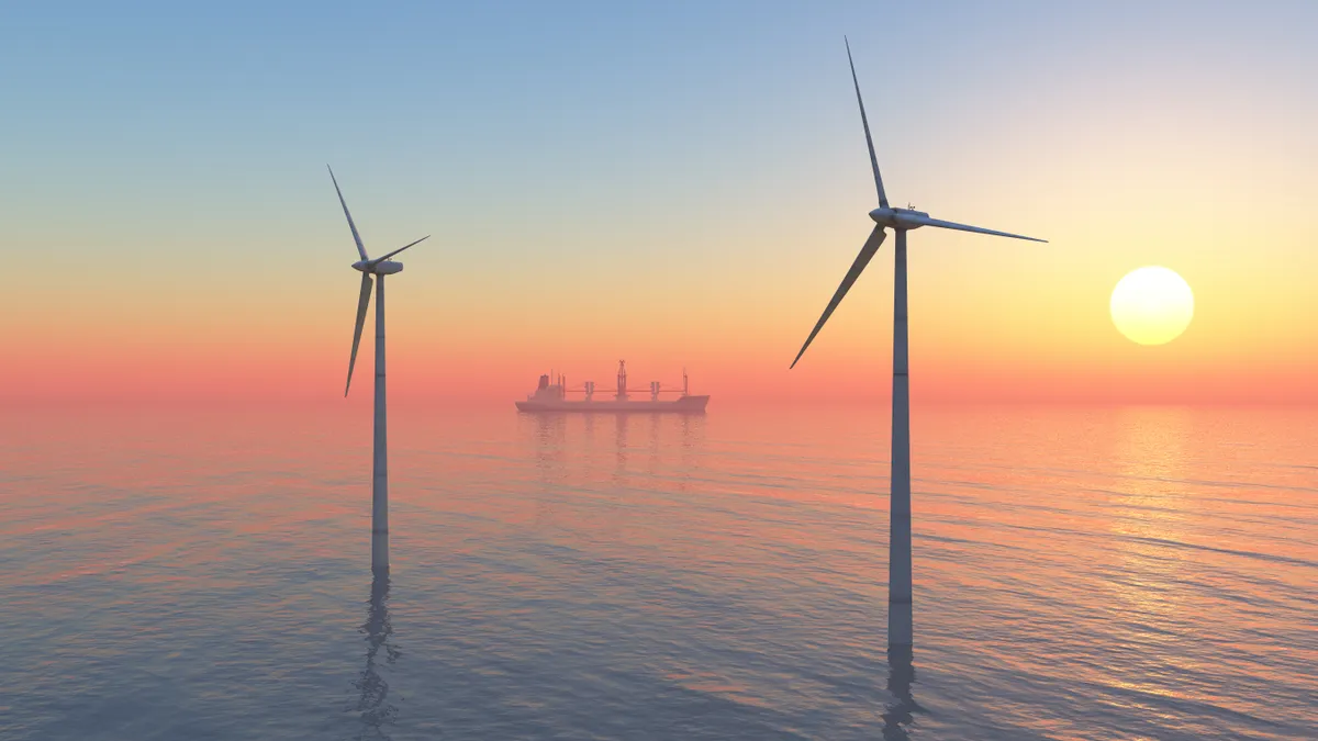 Offshore wind turbines sit in the ocean at sunset with a cargo ship behind them.