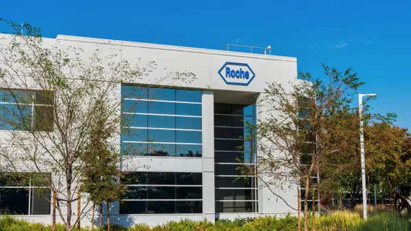 A modern office building at the Roche Diagnostics campus features a sleek glass façade with the Roche logo displayed near the entrance, and the structure is surrounded by landscaped greenery. 