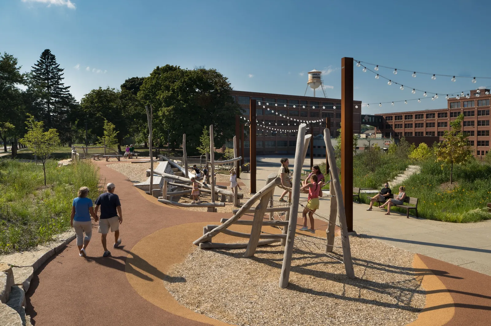 A couple walks holding hands along a path past children playing on wooden play structures.