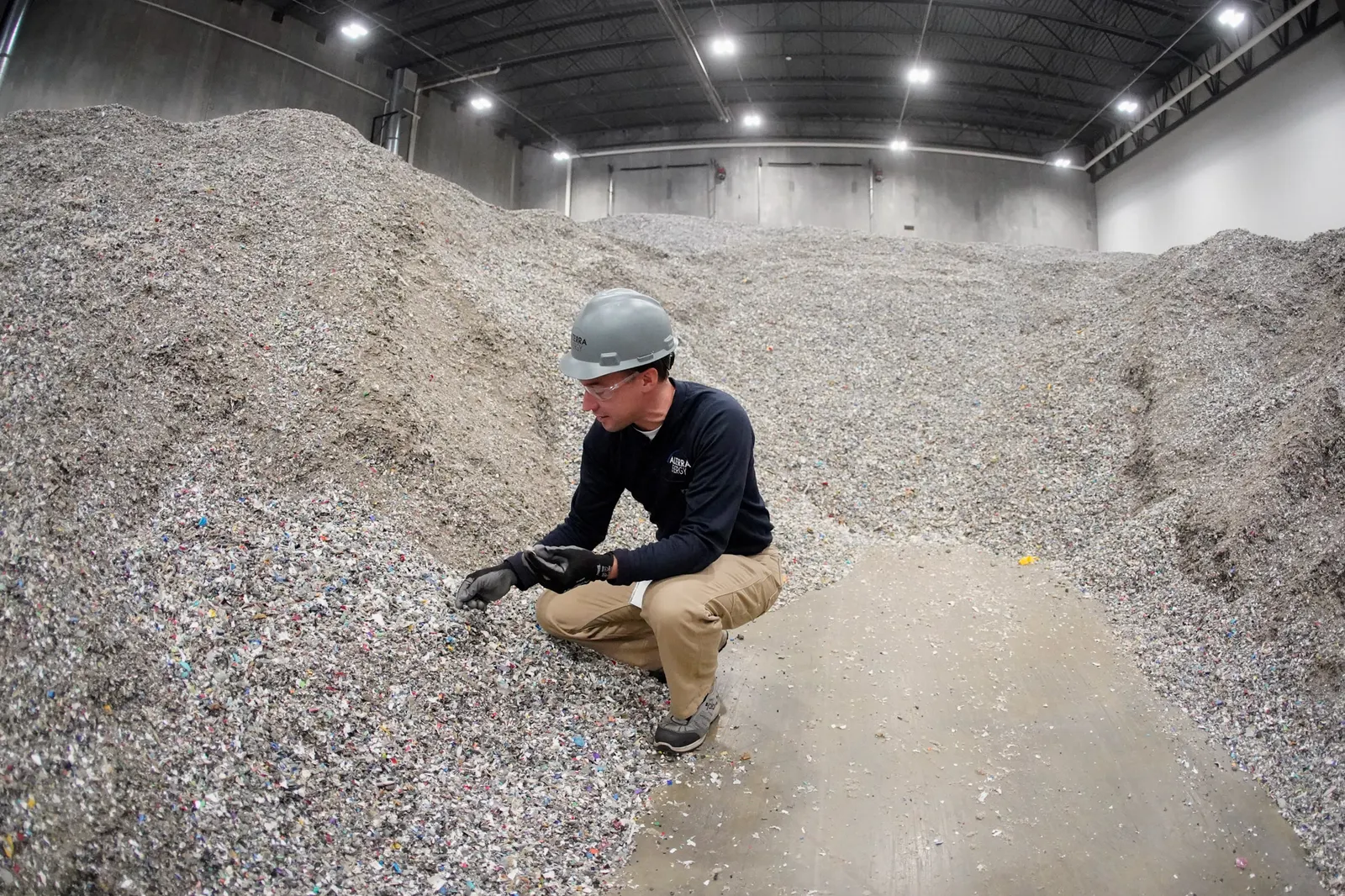 A person wearing a hard hat and safety glasses crouches beside a pile of shredded plastics in a warehouse.