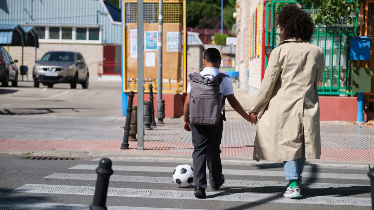 A child crosses the street while holding the hand of their parent. The child is also a kicking a ball to their left.