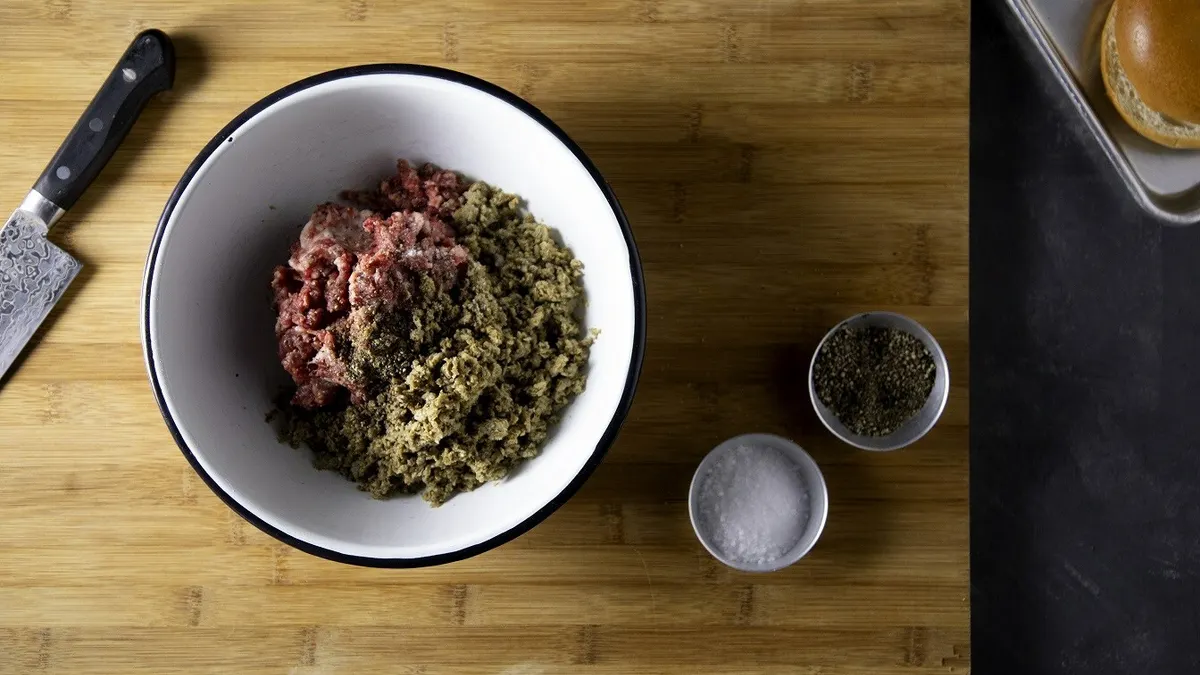 A black-rimmed white bowl with ground beef and Mush Foods' light brown mycelium ingredient sits on a wooden cutting board with a knife to the left and salt and pepper in cups to the right.