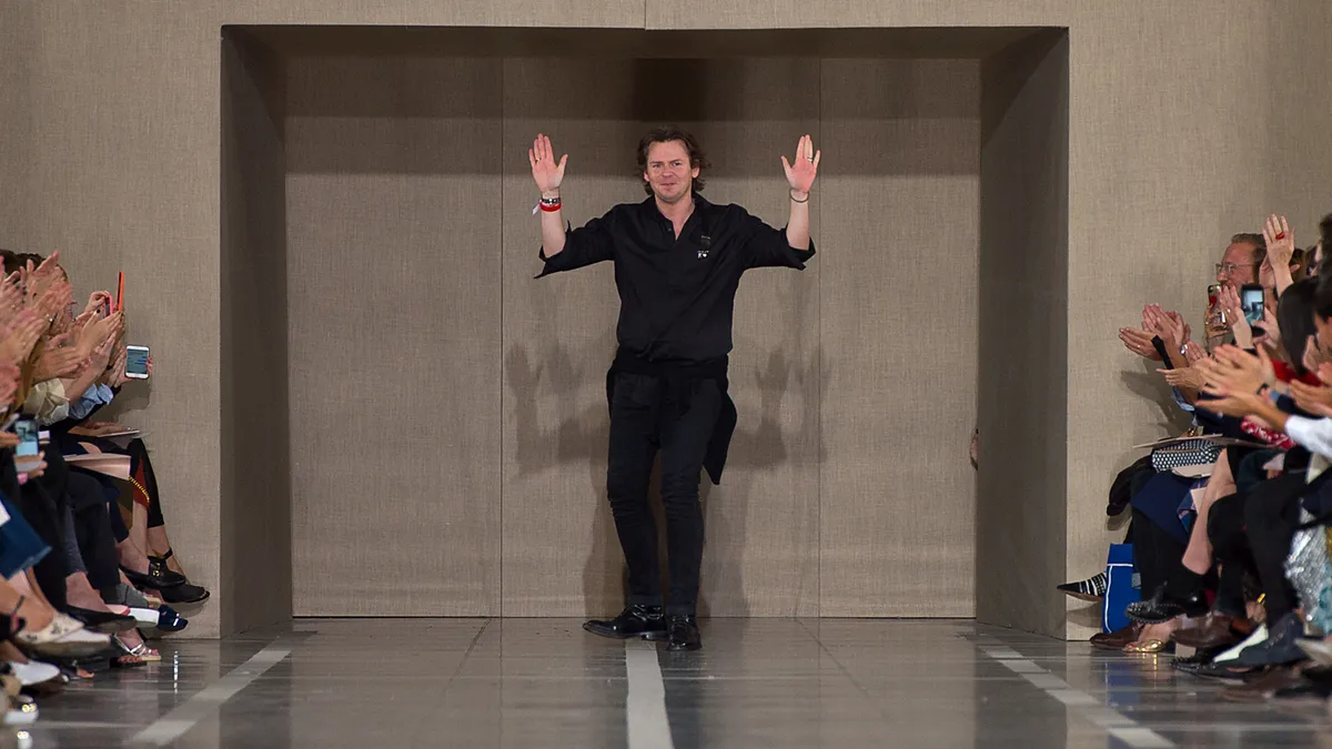 A man in a a black shirt and pants waves with both hands to a seated crowd at the end of a fashion runway.