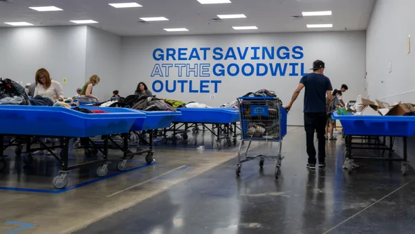 People shopping at a Goodwill outlet store. A person pushes a blue cart down an aisle as others pick through clothing bins.