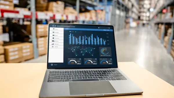A laptop sits on a warehouse desk running a management system dashboard for data analytics, inventory tracking and logistics.