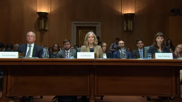 Three people sit at a long table in a hearing room.