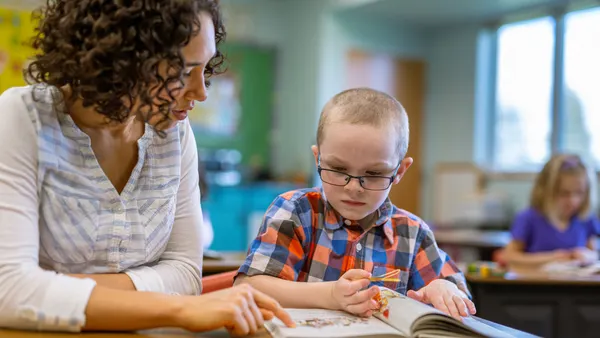 An elementary school student and adult sit at a table in a classroom. They are looking at the inside of a book on the table.