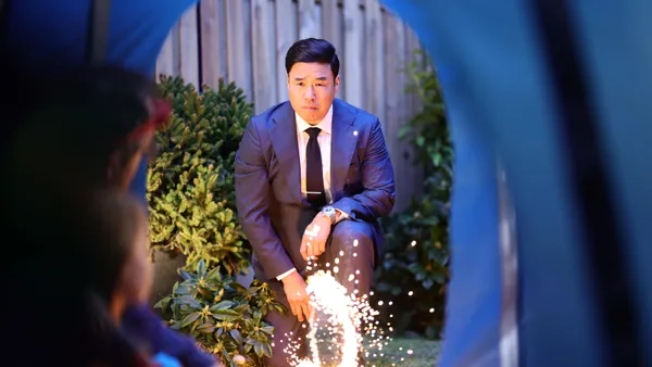 Actor Randall Park kneels in a yard wearing a blue suit as part of Priceline's new ad campaign bringing back the Negotiator character