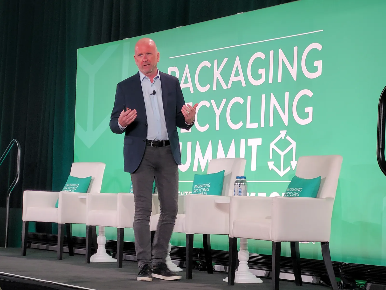 A person walks across a stage while speaking and a green background behind them has the title Packaging Recycling Summit.