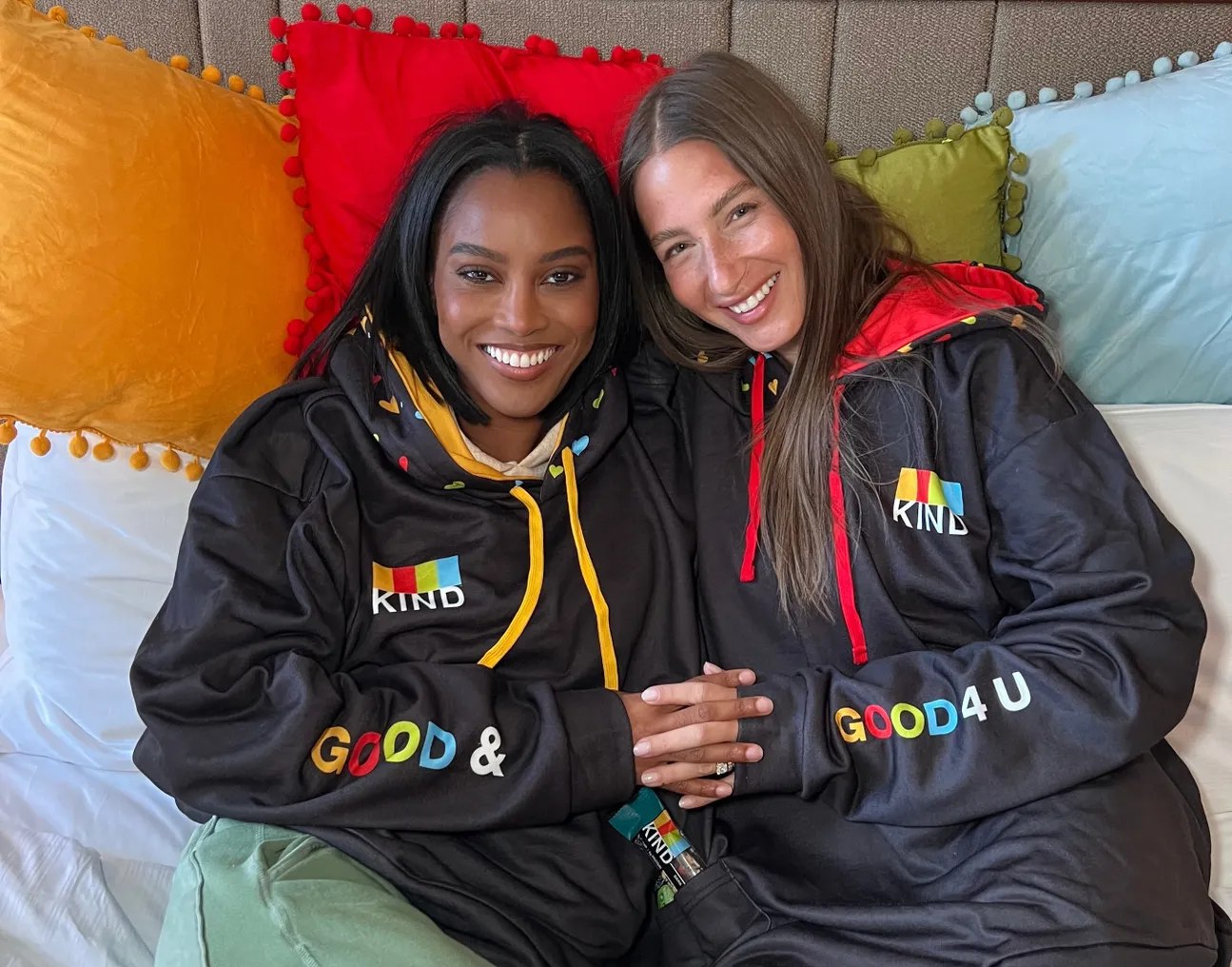 Ciara Miller and Amanda Batula lounge in bed wearing Kind hoodies.