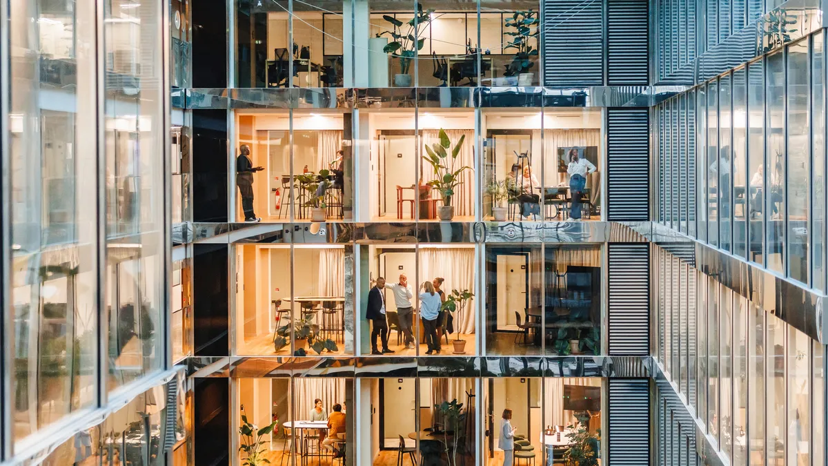 A well-lit office setting shows employees across multiple floors and rooms of the buildings through glass windows.