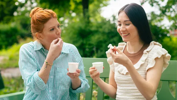 Two people use spoons to eat ice cream out of paper cups while sitting on a bench outdoors.