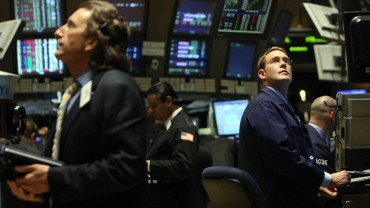 Two men sit at desks looking up at video monitors.