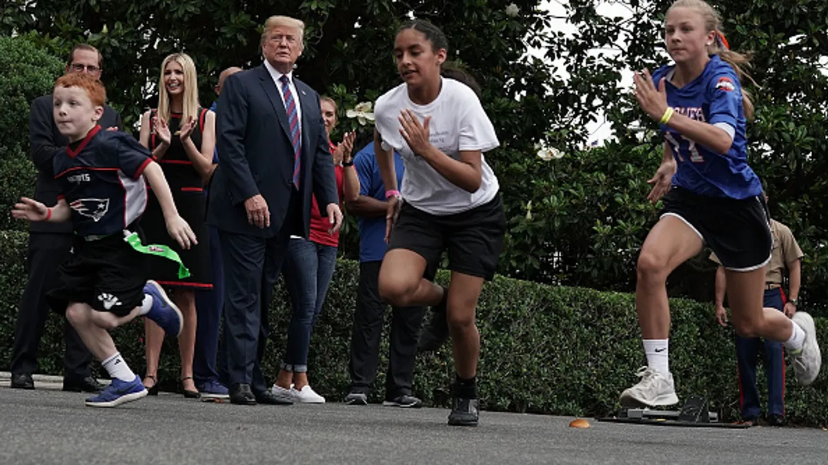 Then-President Donald Trump is pictured outside watching three children run in front of him. Other people are on the sidelines.